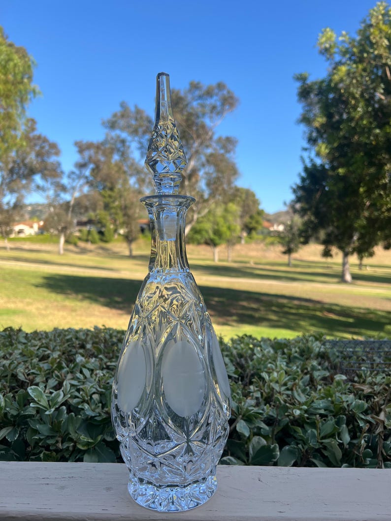 May include: A clear glass decanter with a diamond-cut pattern and a frosted oval design. The stopper is a tall, pointed shape with a diamond pattern. The decanter is displayed outdoors with a green background.