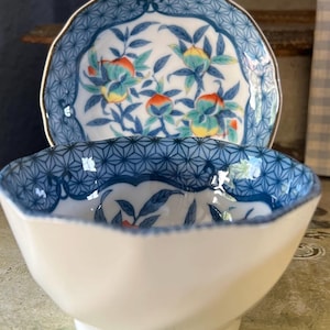 May include: A white ceramic bowl with a blue and white geometric pattern around the rim. The bowl is sitting on top of a matching saucer with a floral design featuring blue leaves and orange fruit.