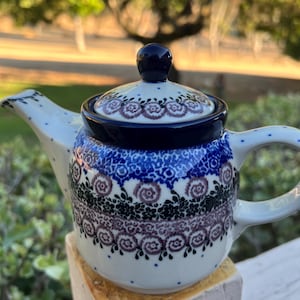 May include: A ceramic teapot with a blue, white, and purple floral pattern. The teapot has a dark blue lid and handle. The spout and handle are white with blue polka dots. The teapot is sitting on a white surface.