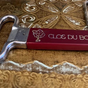 May include: A red bottle opener with a silver handle and the words "CLOS DU BOIS" engraved on the side. A grapevine design is also engraved on the opener.