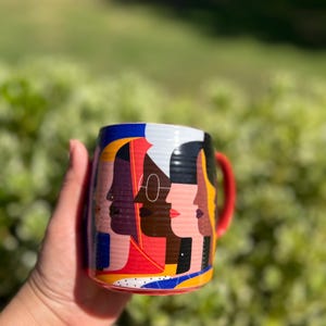 K&ouml;nnte beinhalten: Eine Keramiktasse mit rotem Henkel und einem farbenfrohen Design mit stilisierten Profilen verschiedener Gesichter. Das Design der Tasse enth&auml;lt rote, gelbe, blaue, wei&szlig;e und schwarze Farben. Die Tasse wird in einer Hand vor einem verschwommenen gr&uuml;nen Hintergrund gehalten.