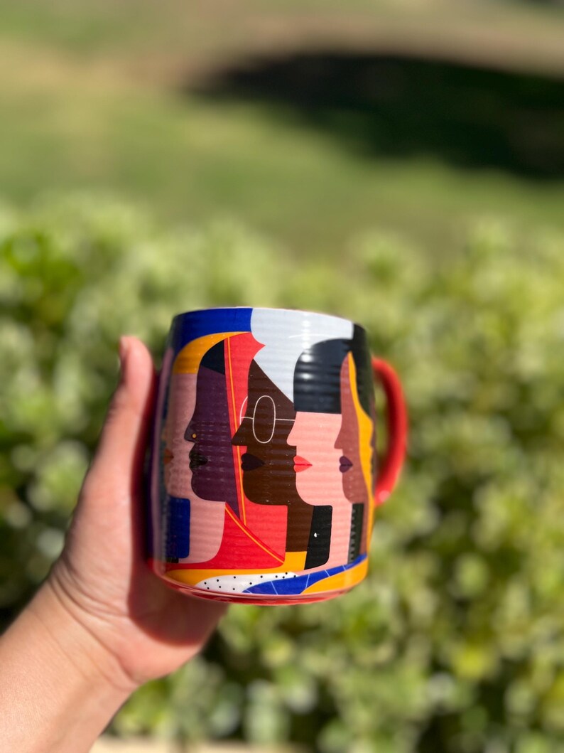 K&ouml;nnte beinhalten: Eine Keramiktasse mit rotem Henkel und einem farbenfrohen Design mit stilisierten Profilen verschiedener Gesichter. Das Design der Tasse enth&auml;lt rote, gelbe, blaue, wei&szlig;e und schwarze Farben. Die Tasse wird in einer Hand vor einem verschwommenen gr&uuml;nen Hintergrund gehalten.