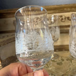 May include: Clear wine glass with etched winter scene of trees and snow. The glass is held in a hand, with other glasses visible in the background. The etched design is white.