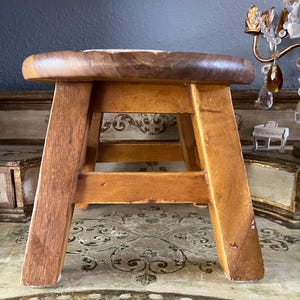 May include: A small wooden stool with a round top and four angled legs. The stool features a natural light brown wood grain finish with visible texture and signs of wear. The structure consists of a circular seat supported by four sturdy legs connected by horizontal crossbars for stability.