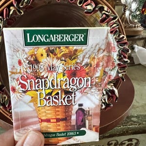 May include: A Longaberger 1998 May Series Snapdragon Basket card is held in front of a decorative basket. The card features a watercolor illustration of a woven basket filled with colorful flowers. The basket has a brown rim with a decorative ribbon.