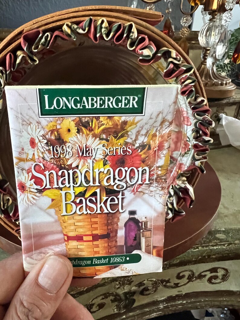 May include: A Longaberger 1998 May Series Snapdragon Basket card is held in front of a decorative basket. The card features a watercolor illustration of a woven basket filled with colorful flowers. The basket has a brown rim with a decorative ribbon.