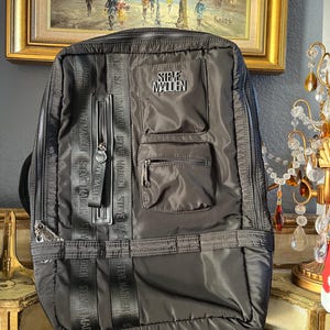 May include: Black Steve Madden backpack with multiple zippered compartments and the brand name prominently displayed. The backpack features vertical and horizontal straps with the Steve Madden logo, and a top handle.