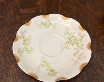 May include: A white porcelain saucer with a scalloped edge and a gold trim. The saucer is decorated with a delicate floral pattern in shades of pink and green.