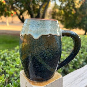 May include: A handcrafted ceramic mug with a unique design. The mug features a dark blue and green glaze with a light blue rim. The handle is a matching dark blue. The mug is sitting on a white wooden post, with a blurred green background.
