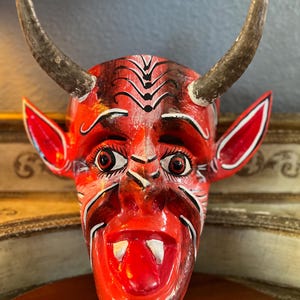 May include: A red wooden mask with horns, painted white and black details. The mask has a wide open mouth with a red tongue and white teeth.