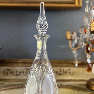 May include: Clear crystal glass decanter with a diamond-cut pattern and a pointed stopper. The decanter has a long, slender neck and a bulbous body with two frosted oval accents. The stopper is intricately cut with a floral design.