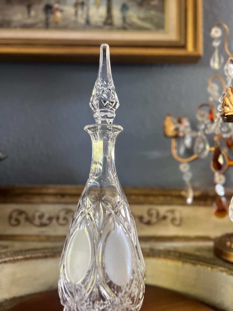May include: Clear crystal glass decanter with a diamond-cut pattern and a pointed stopper. The decanter has a long, slender neck and a bulbous body with two frosted oval accents. The stopper is intricately cut with a floral design.