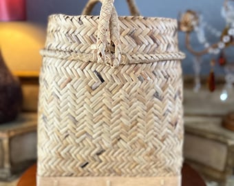 May include: A woven natural fiber basket with a handle. The basket is light brown and has a woven pattern. The basket is sitting on a wooden surface.