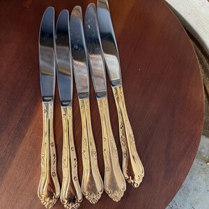 May include: Five silver dinner knives with gold-plated handles. The handles have an ornate floral design.