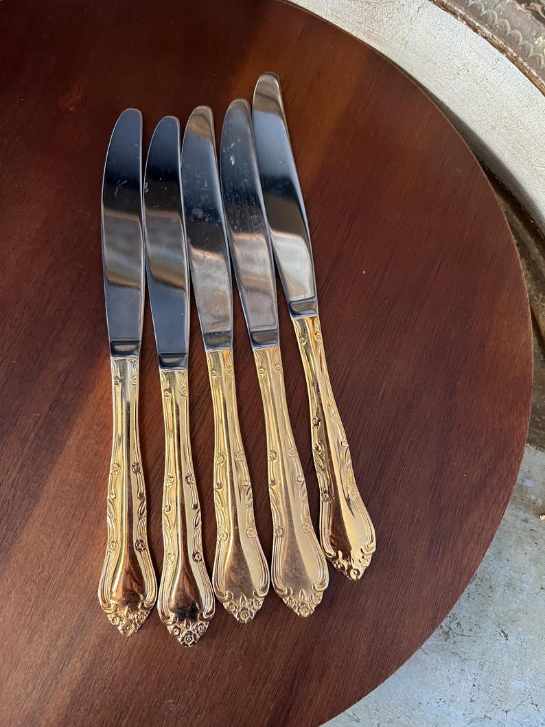 May include: Five silver dinner knives with gold-plated handles. The handles have an ornate floral design.