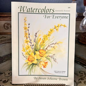 May include: A book titled "Watercolors For Everyone" by Susan Scheewe Brown. The cover features a watercolor painting of yellow daffodils and other spring flowers. The book is propped up on a decorative stand.
