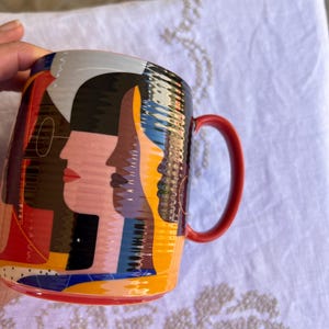 K&ouml;nnte beinhalten: Eine Keramiktasse mit einem lebendigen Design, das stilisierte Gesichter in Rosa, Schwarz, Gelb und Blau zeigt. Die Tasse hat einen roten Henkel und einen roten Rand am Boden. Das Design umschlie&szlig;t die Tasse.
