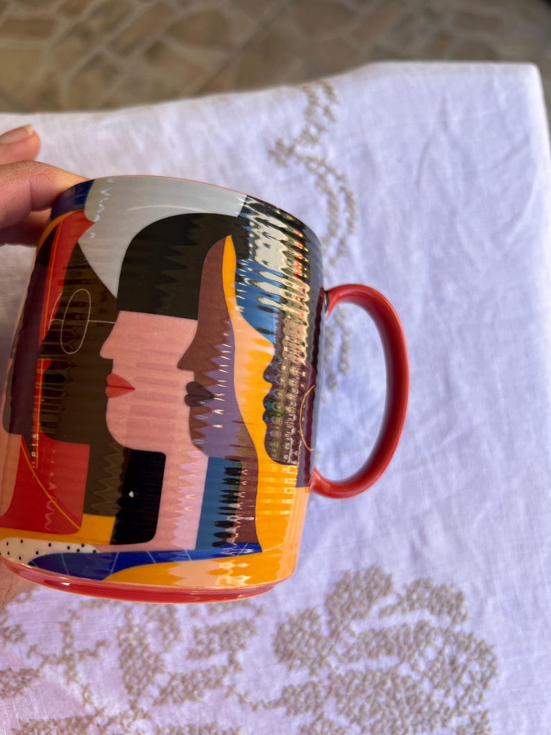 K&ouml;nnte beinhalten: Eine Keramiktasse mit einem lebendigen Design, das stilisierte Gesichter in Rosa, Schwarz, Gelb und Blau zeigt. Die Tasse hat einen roten Henkel und einen roten Rand am Boden. Das Design umschlie&szlig;t die Tasse.