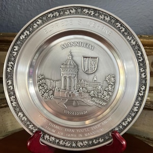May include: A silver-colored decorative plate with embossed details. The plate features the text "ELFRIEDE S. GEHRKE" and "MANNHEIM", along with an image of a building and the words "WASSERTURM". The plate has a grape vine border and the text "MORE THANKS THAN YOU'LL EVER KNOW FROM THE GUYS IN MANNHEIM".