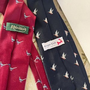 May include: Two neckties with a bird pattern. One is red with a pheasant pattern and the other is navy blue with a duck pattern. The red tie has a "Resilio" label and the navy blue tie has a "Cape Cod Neckwear" label.