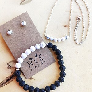 Op de afbeelding: Een sieradenset met een zilveren ketting en armband met wit-zwarte gemarmerde kralen, en een zwarte lavasteenkralenarmband. Bijpassende oorknopjes worden op een bruine kaart getoond. Het Rye Jewelry-logo is zichtbaar.