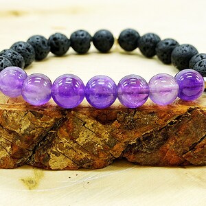 May include: A beaded bracelet featuring alternating black lava rock and vibrant purple amethyst beads. The bracelet is strung on a stretch cord and includes gold-colored spacer beads. The bracelet is displayed on a piece of wood.