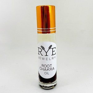 Puede incluir: Un frasco roll-on de vidrio transparente con tapa dorada. El frasco tiene una etiqueta blanca con el texto "RYE JEWELRY" y "ROOT CHAKRA OIL". El aceite en su interior es de color ámbar claro con un sedimento marrón oscuro en el fondo.