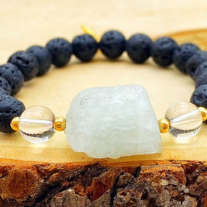 Peut inclure: Un bracelet composé de perles de roche volcanique noire, d'une grosse pierre bleu-vert clair et de perles transparentes avec des accents dorés. Le bracelet est monté sur un cordon élastique et est présenté sur une surface en bois.