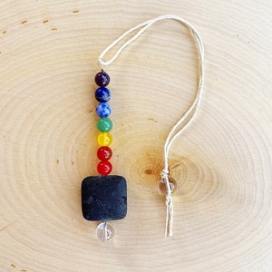 Puede incluir: Un adorno colgante con temática de chakras que presenta una cadena de cuentas coloridas en un espectro de arcoíris, una piedra cuadrada negra y una esfera de cristal transparente. El adorno está suspendido por un cordón de color natural.