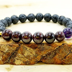 May include: A bracelet with black lava rock beads, deep red garnet beads, and a single purple amethyst bead. The bracelet has gold-coloured spacers. The beads are approximately 0.8 cm in diameter.