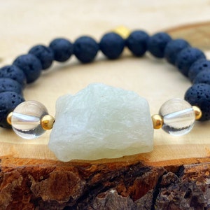 Op de afbeelding: Een armband met zwarte lavasteen kralen, een grote lichtgroene steen, heldere kralen en goudkleurige accenten. De armband wordt gepresenteerd op een houten oppervlak.