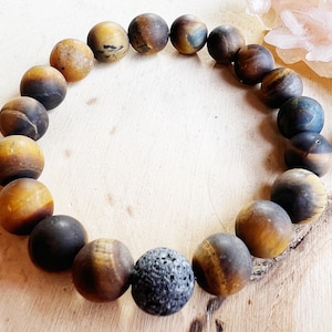 Op de afbeelding: Een armband gemaakt van ronde tijgeroog- en lavasteen kralen. De tijgeroogkralen hebben een mix van bruine, gouden en zwarte tinten. De lavasteenkraal is donkergrijs en poreus. De armband wordt gepresenteerd op een licht houten oppervlak.