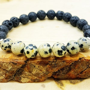 Könnte beinhalten: Ein Armband mit schwarzen Lavasteinperlen und gesprenkelten Dalmatiner-Jaspis-Perlen. Das Armband ist auf einem elastischen Band aufgefädelt. Das Schmuckstück wird auf einer Holzoberfläche präsentiert.