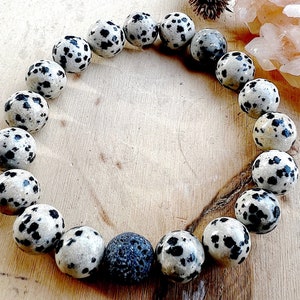 May include: A collection of round, speckled stones arranged in a circular shape. The stones are primarily off-white with black spots, resembling dalmatian markings. One stone is a dark gray color. The arrangement is on a light brown wooden surface.