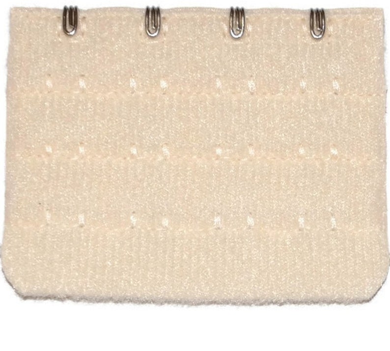 May include: A white bra extender with four metal hooks and eyes. The extender has a textured surface and is designed to adjust the length of a bra strap.
