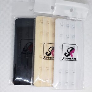 Peut inclure: Trois extensions de soutien-gorge noires, beiges et blanches, chacune emball&eacute;e dans un sac en plastique transparent. Chaque extension comporte trois rang&eacute;es de crochets et d'agrafes. Le logo RorosArt est visible.