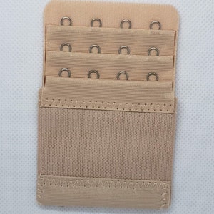 May include: A beige bra extender with three rows of hooks and eyes. Each row has three hooks and eyes.