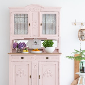 Könnte beinhalten: Ein hellrosa Holzschrank mit zwei oberen Glastüren, zwei Schubladen und zwei unteren Schranktüren mit geschnitzten Blumendetails. Der Schrank verfügt über eine offene Ablagefläche in der Mitte mit einem Spiegel und Messingbeschlägen.