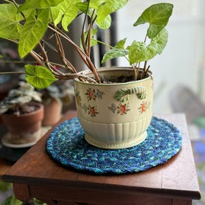 May include: A cream-colored ceramic planter with a fluted base and two side handles, decorated with small orange floral and blue leaf patterns. The planter holds a green Syngonium plant and sits on a round, braided blue and teal fabric mat.