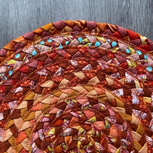 Könnte beinhalten: Ein runder geflochtener Teppich mit einem kreisförmigen Muster in Orange-, Braun- und Rosatönen. Der Teppich besteht aus Stoffresten, die zu einem kreisförmigen Muster zusammengenäht wurden.