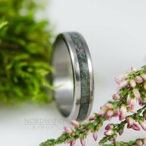 Peut inclure: Une bague en titane argentée avec un incrustation de pierre verte. La bague est placée sur un fond de mousse verte et de fleurs roses.