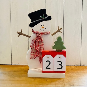 May include: A white wooden snowman with a black top hat, red and white plaid scarf, and a green tree. The snowman is holding a red wooden block with white numbers 23.