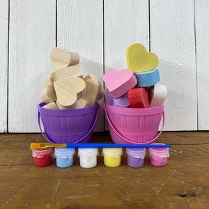 May include: Two buckets filled with wooden heart shapes, one purple bucket with natural wood hearts and one pink bucket with painted hearts in pink, yellow, blue and red.  A paintbrush and six small paint bottles in red, blue, white, yellow, purple and pink are in front of the buckets.