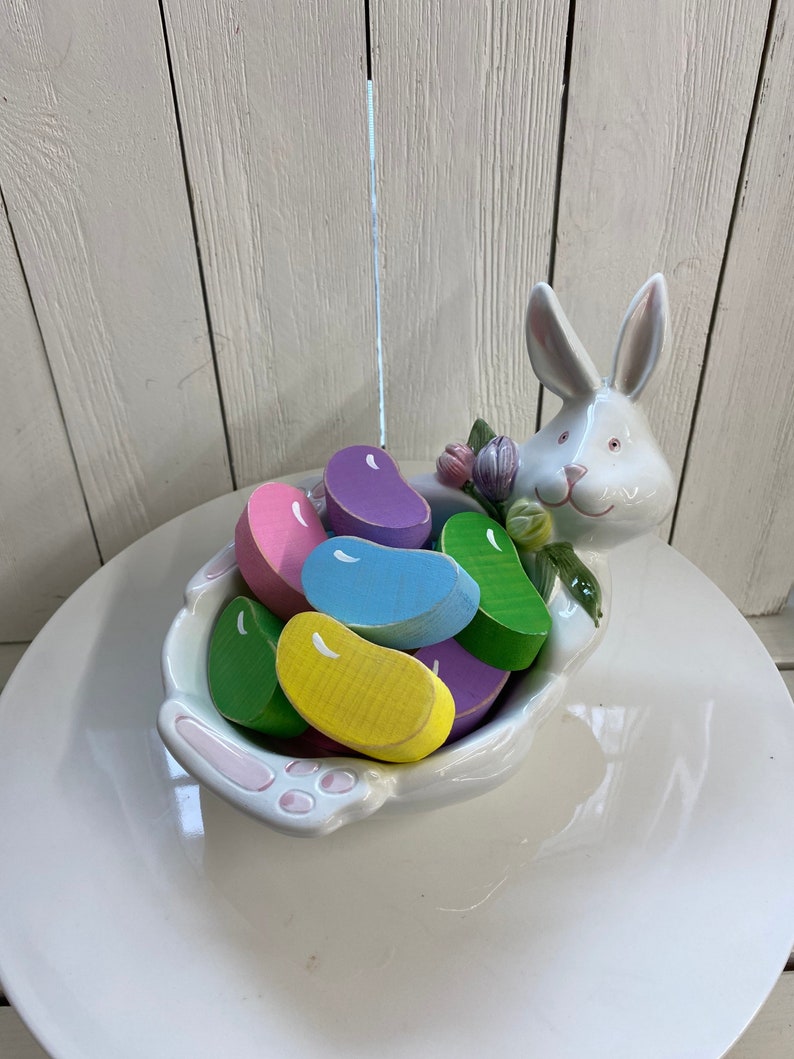 May include: White ceramic bunny dish with a pink rim and floral accents. The dish is filled with colorful wooden jelly bean shaped candies.