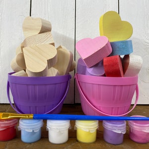 May include: A craft kit with two buckets filled with wooden heart shapes, one bucket is purple and the other is pink. The buckets are on a wooden surface with six small paint jars in front of them. The paint jars are red, blue, white, yellow, purple, and pink. A blue paintbrush is lying on the surface in front of the paint jars.