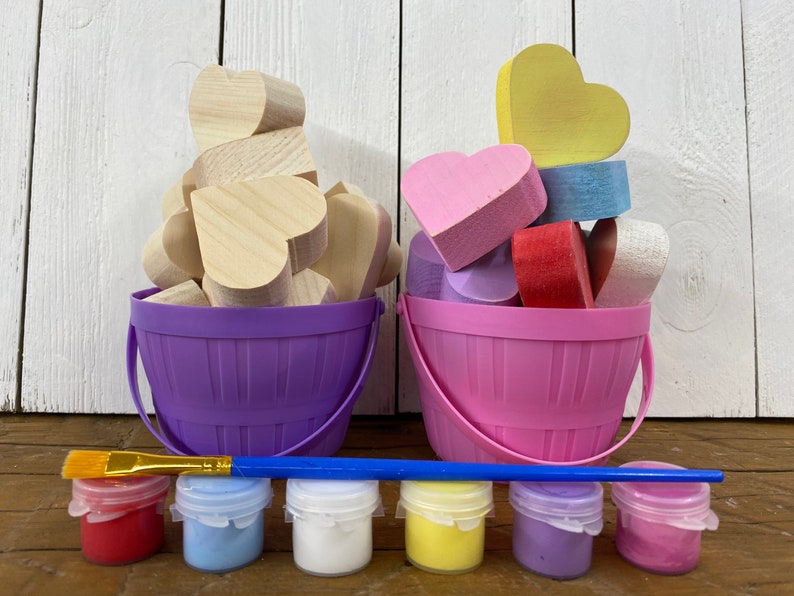 May include: A craft kit with two buckets filled with wooden heart shapes, one bucket is purple and the other is pink. The buckets are on a wooden surface with six small paint jars in front of them. The paint jars are red, blue, white, yellow, purple, and pink. A blue paintbrush is lying on the surface in front of the paint jars.