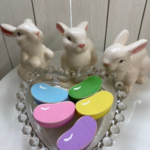 May include: Five wooden jelly bean shaped ornaments in a heart-shaped glass dish. The jelly beans are painted in pastel colors: blue, pink, yellow, green, and purple. Three white ceramic bunnies surround the dish.