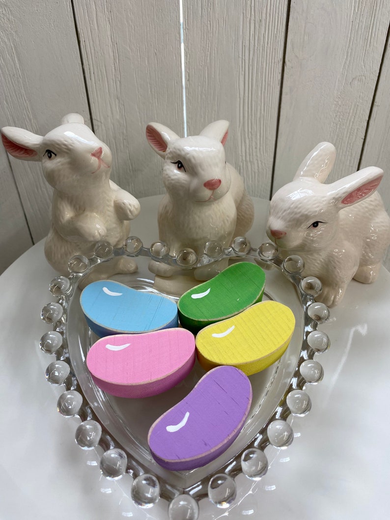 May include: Five wooden jelly bean shaped ornaments in a heart-shaped glass dish. The jelly beans are painted in pastel colors: blue, pink, yellow, green, and purple. Three white ceramic bunnies surround the dish.