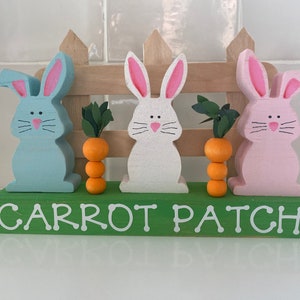 May include: Three wooden bunny figurines painted pink, blue, and white stand in front of a wooden fence with a green base that reads "Carrot Patch". Each bunny has a stack of three orange carrot-shaped decorations in front of it.