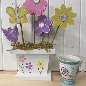 May include: A white planter with a wooden base, decorated with painted flowers, holds a bouquet of wooden flowers. The flowers are painted in shades of pink, purple, yellow, and green. A butterfly is perched on one of the stems. A white teacup with a floral design sits on a wooden surface.
